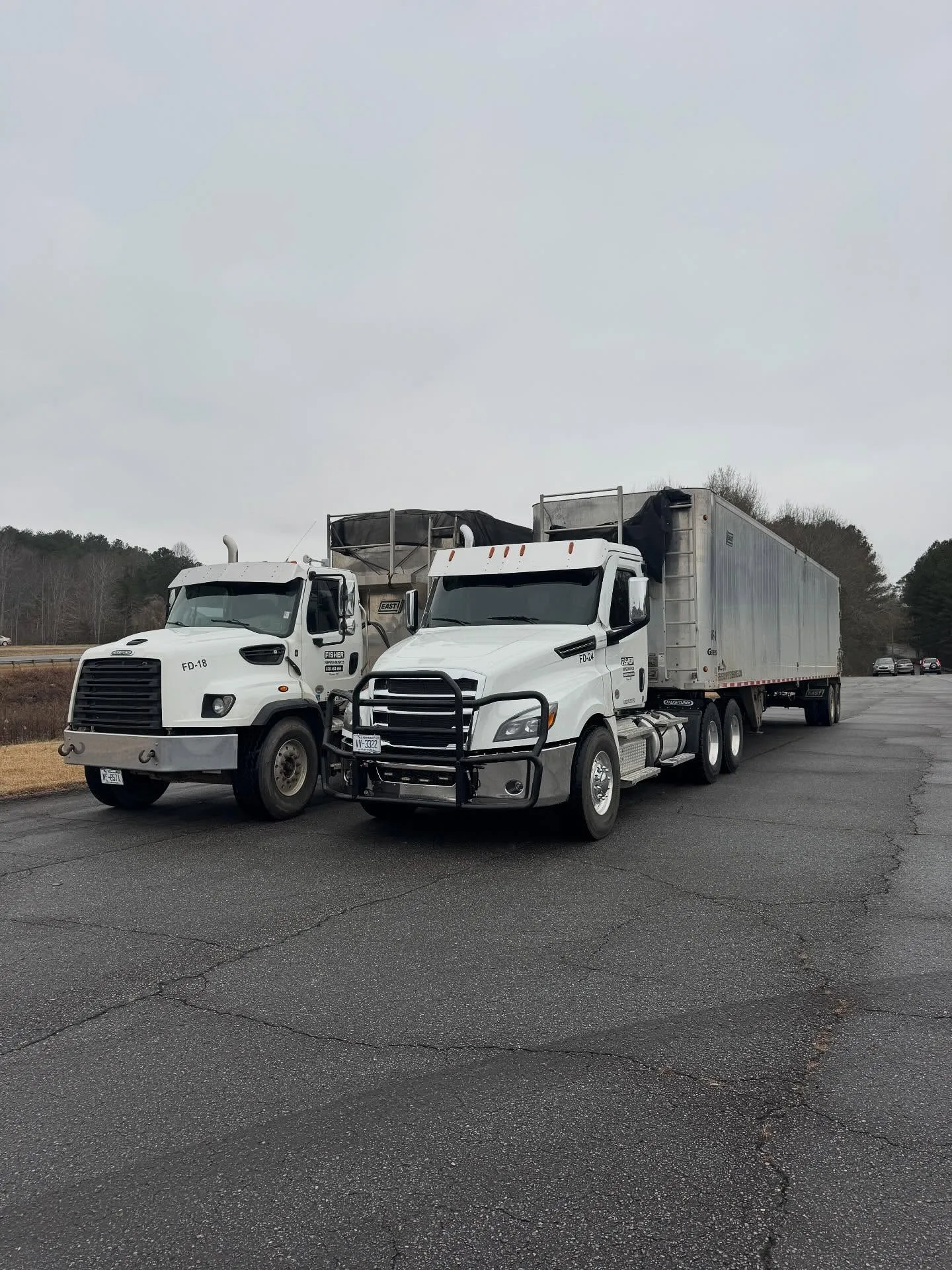 Two large Freightliner trucks parked on a road surrounded by trees.
