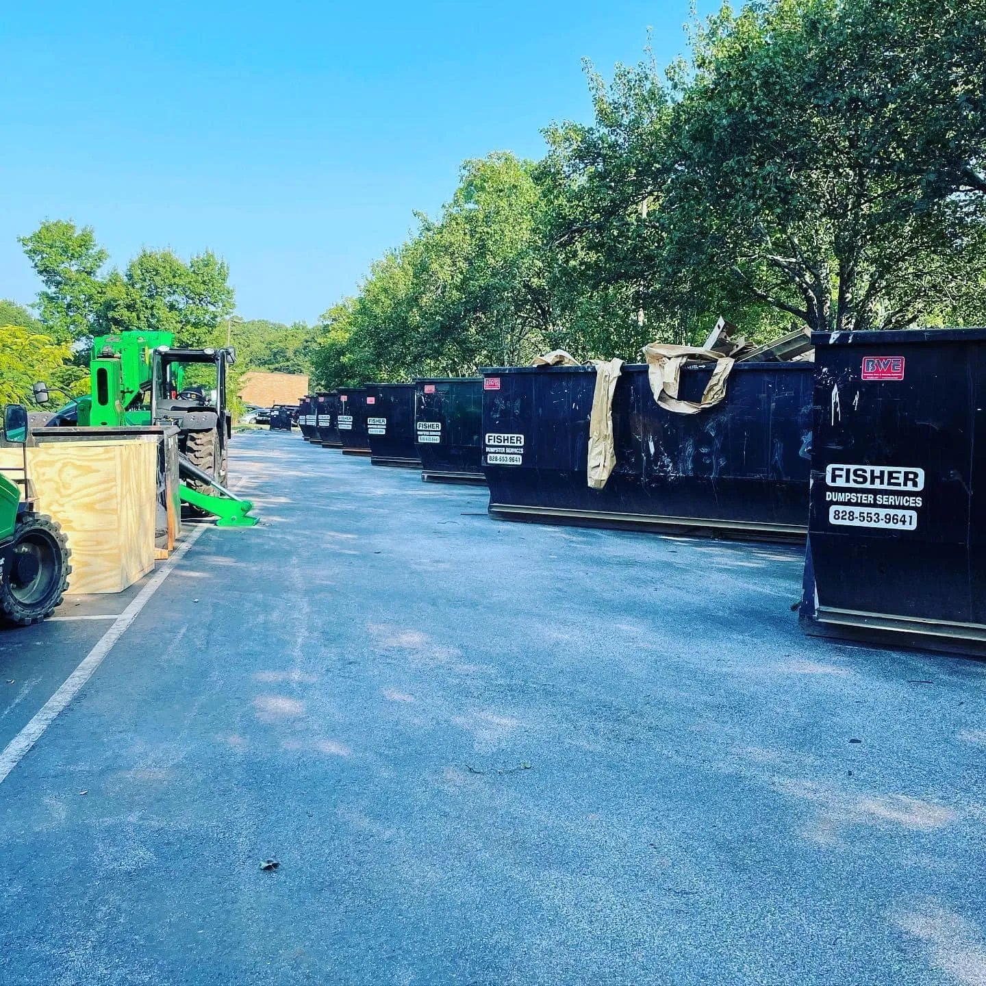 Row of dark dumpsters along a paved road with green construction equipment nearby.